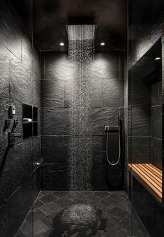 a shower head in the middle of a black tiled bathroom with wooden bench under it