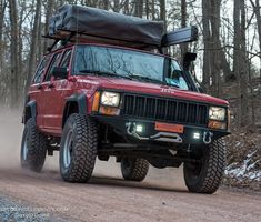 a red jeep driving down a dirt road in the woods with its hood up and lights on