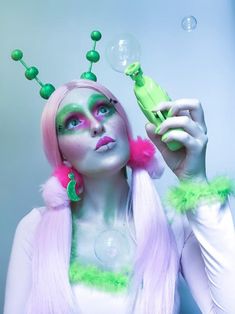 a woman with pink hair and green makeup is blowing bubbles in front of her face