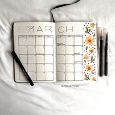 an open planner with flowers on it next to two markers and a pen, sitting on top of a white sheet