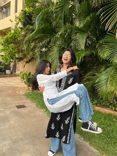 two women standing next to each other in front of palm trees and bushes, one carrying the woman on her back