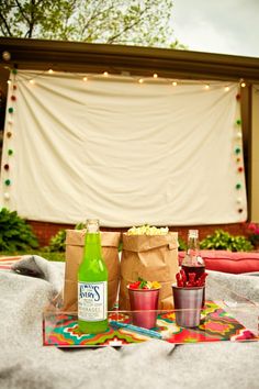 an outdoor picnic with drinks and snacks on the table in front of a white curtain