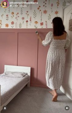 a woman standing in front of a pink painted wall next to a bed and dresser