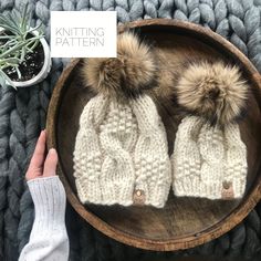 two knitted hats with pom - poms sitting on top of a wooden bowl