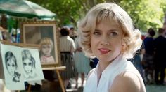 a woman with blonde hair standing in front of an easel and some pictures on display