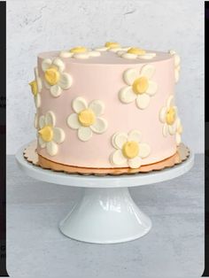 a pink cake with white and yellow flowers on it, sitting on a pedestal in front of a wall