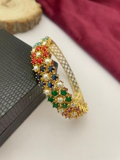 This stunning handcrafted bangle bracelet features a vibrant combination of multicolor stones and pearls set in a luxurious gold-plated design. Perfect for adding a splash of color to any outfit, this bangle is ideal for bridal jewelry or festive occasions such as weddings, Diwali, or family celebrations. The intricate detailing and adjustable fit make this bracelet both stylish and comfortable, ensuring it can be worn for any event with ease. This ethnic cuff bangle is a versatile accessory that can complement both traditional and contemporary outfits, making it a must-have for jewelry lovers. Whether you're looking for a standout piece for your bridal collection or a unique gift for someone special, this handcrafted multicolor bangle is sure to impress. Elegant Multicolor Beaded Bracelets For Festive Occasions, Elegant Multicolor Multi-stone Cuff Bracelet, Elegant Multicolor Beaded Bangle Bracelet, Elegant Multicolor Gold Plated Bracelets, Elegant Multicolor Gold-plated Bracelets, Multicolor Gold Plated Bracelet, Multicolor Jeweled Bangle, Elegant Multicolor Festive Bracelets, Multicolor Beaded Bangle Bracelets For Wedding