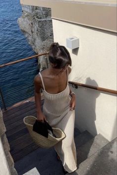 a woman in a white dress is walking up stairs next to the water and holding a hat