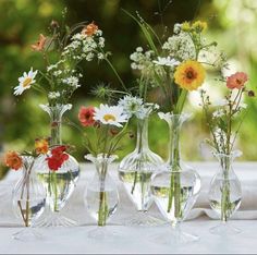 four vases with flowers in them sitting on a white table cloth outside near some trees