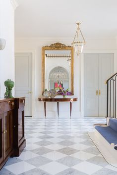 a hallway with white walls and checkered tile flooring, gold framed mirror on the wall