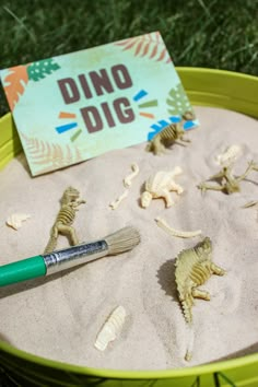 a green bucket filled with sand and toy dinosaurs next to a sign that says dino dig