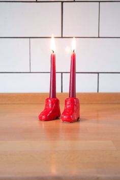 two candles with red shoes on them sitting on a wooden table in front of a white brick wall