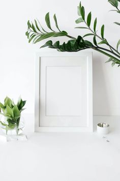 a white frame sitting on top of a table next to a vase filled with green leaves