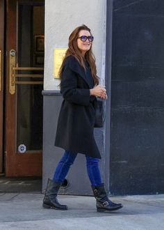 a woman in black coat and blue jeans walking on sidewalk next to building with door