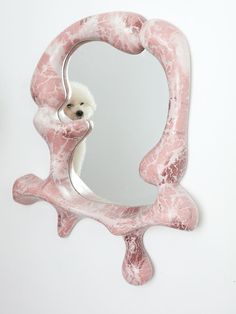 a small white dog is peeking out of a pink marbled wall mounted vanity mirror