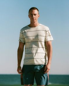 a man standing in front of the ocean with his hands in his pockets, wearing shorts and a striped t - shirt