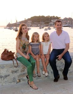 a family sitting on a stone wall next to the water with boats in the background