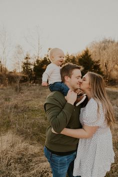 a man holding a baby while standing next to a woman in a field with trees