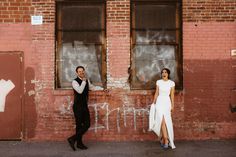 a man and woman standing next to each other in front of a brick wall with graffiti on it