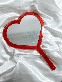 a red heart shaped mirror sitting on top of a white satin covered bed sheet,