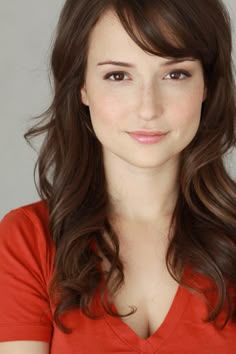 a woman with long brown hair wearing a red shirt and smiling at the camera while standing in front of a gray background