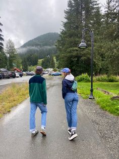 two people are walking down the road in front of some trees and cars on a cloudy day