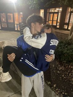 two people are hugging each other on the sidewalk at night, one is wearing a blue and white jacket