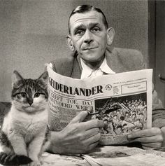 a man sitting at a table reading a newspaper with a cat on his lap next to him