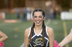 a girl with braids smiles while standing in front of other cheerleaders on the field