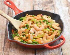 a skillet filled with shrimp, peas and carrots on a wooden table next to a red spatula