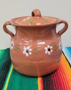 a brown pot with flowers painted on it sitting on a colorful table cloth next to a white wall