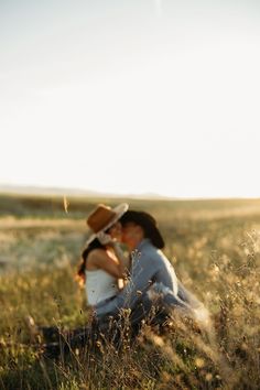 a man and woman sitting in the grass with a cowboy hat on their head looking at each other