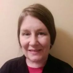 a woman wearing a red shirt and black jacket smiles at the camera while standing in front of a wall