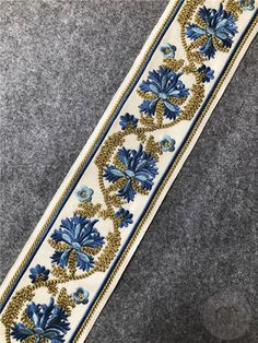 a white and blue ribbon with gold trimmings on grey fabric, in the shape of an ornament