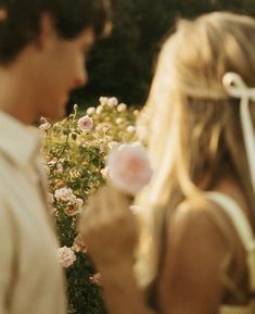 a man and woman standing next to each other in front of flowers with one blowing the dandelion