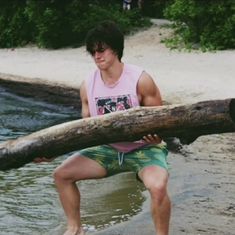 a man holding a large log in his hand on top of a beach next to water