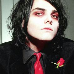 a man with long black hair wearing a red tie and rose in his lapel