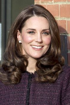 a woman with long brown hair and wearing a purple sweater smiles at the camera while standing in front of a brick wall