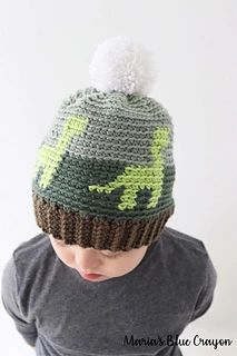 a young boy wearing a knitted dinosaur hat with pom - pom on top