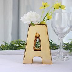 a gold letter vase with flowers in it on a table next to wine glasses and greenery