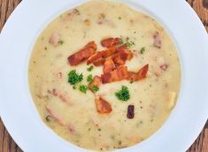 a white bowl filled with soup and bacon on top of a wooden table next to a spoon