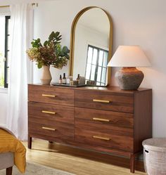 a bedroom with a dresser, mirror and lamp on it's side table in front of a window