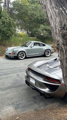 two sports cars parked next to each other in a parking lot with trees and bushes