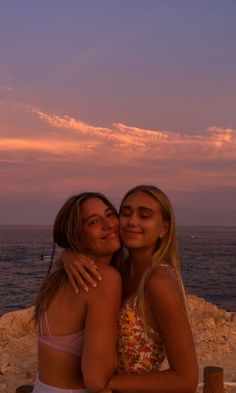 two young women hugging each other on the beach at sunset, with water in the background