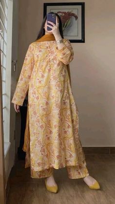 The image shows a person taking a mirror selfie, showcasing their traditional outfit. The attire consists of a loose-fitting, long-sleeved kameez with a light yellow and orange floral pattern, paired with matching straight-cut trousers. The outfit has a soft, summery feel, featuring light and bright colors. The person is also wearing a mustard-colored dupatta, draped casually over one shoulder. They have accessorized with silver bangles on their wrist and yellow pointed flats that complement the outfit. A framed floral painting can be seen in the background, adding a touch of elegance to the setting. A Mirror Selfie, Kameez Designs, Traditional Outfit