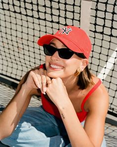 a woman sitting on the ground in front of a tennis net wearing a red hat and sunglasses