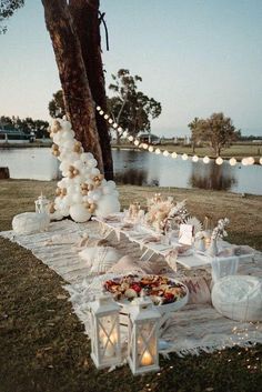 an outdoor table set up for a party with balloons and lights on the grass next to a lake