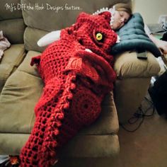 a child is sleeping on a couch with a crocheted dragon blanket over it