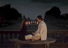 a man and woman sitting on a bench in front of trees at night, talking to each other