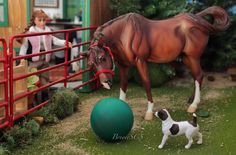 a toy horse standing next to a green ball in front of a red fence with a small white and black dog